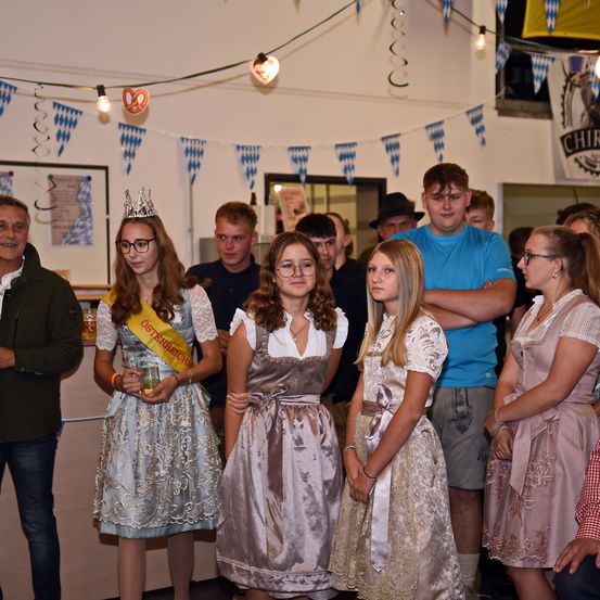Eine Gruppe von Menschen in traditioneller deutscher Kleidung, einschließlich einer Krone, steht in einem für ein Fest dekorierten Raum. Lichterketten und Wimpel sind sichtbar.