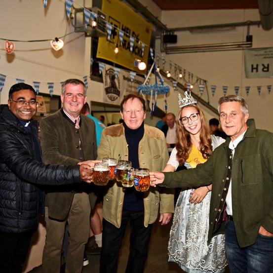 Fünf Personen bei einem festlichen Ereignis, zwei halten Bierkrügen in den Händen und lächeln. Eine Frau in traditionellem Kleid und Krone. Ein Banner mit Text hängt im Hintergrund.