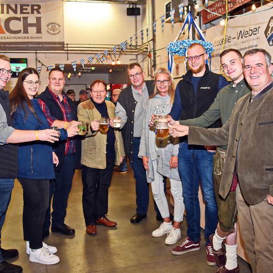 Eine Gruppe von Erwachsenen, die lächeln und Gläser mit Bier halten, stehen bei einer Bierveranstaltung zusammen. Sie befinden sich in einem Raum mit Bannern, einer festlichen Atmosphäre und einigen Personen im Hintergrund.