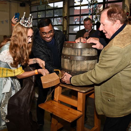 Eine Gruppe von Menschen steht um einen Holzfass herum. Eine Frau mit Krone hält einen Hammer, während ein Mann lächelt und sie ansieht. Ein anderer Mann hält ein Mikrofon und steht in der Nähe. Die Szene findet in einem Innenbereich mit Glasfenstern statt.