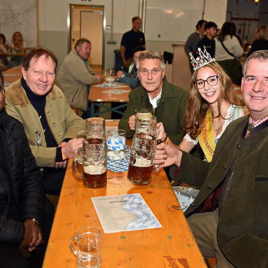 Eine Gruppe von Männern und einer Frau in traditioneller Kleidung sitzen an einem Tisch und heben Biergläser. Die Frau trägt eine Krone. Hinter ihnen sitzen andere Personen an Tischen. Der Raum ist gut beleuchtet.