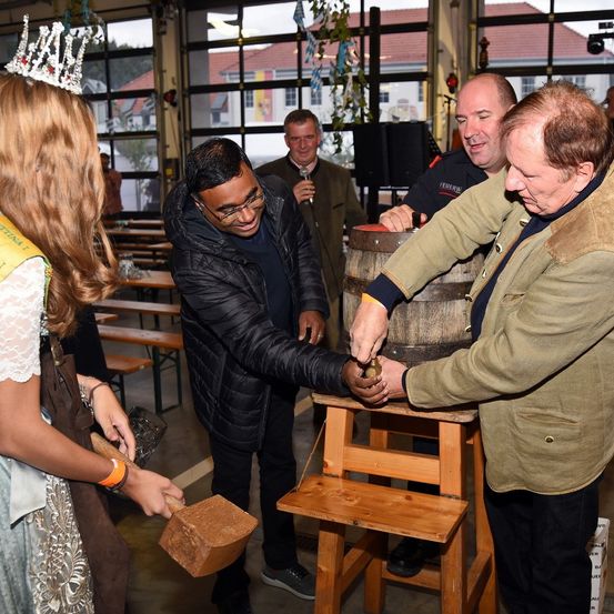 Eine Gruppe von Erwachsenen und einem Kind befindet sich in einer Halle, wo zwei Männer kurz davor sind, mit einem Hammer auf einen Fass zu klopfen. Eine Frau mit Krone hält einen Hammer in den Händen.