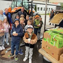 Eine Gruppe von Kindern und Erwachsenen steht vor einem großen orangefarbenen Traktor. Sie halten Tassen und scheinen an einer Veranstaltung teilzunehmen. Hinter ihnen bedient ein Mann eine Maschine, die Behälter mit Lebensmitteln füllt.