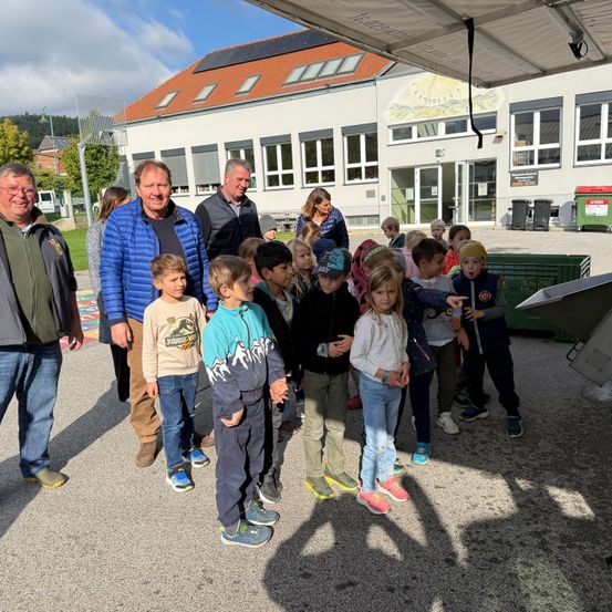 Eine Gruppe von Erwachsenen und Kindern steht vor einem Gebäude mit Solarpaneelen auf dem Dach und schaut etwas an.