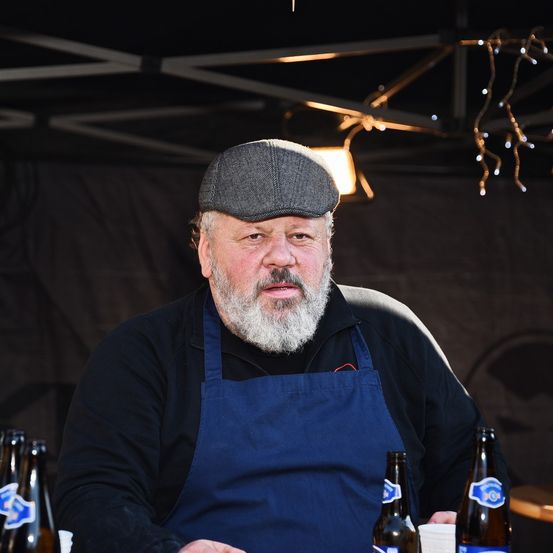 Ein älterer Mann mit Bart und Schnurrbart, der eine Schirmmütze und eine Schürze trägt, sitzt in einem Zelt und bereitet möglicherweise Essen oder Getränke zu. Flaschen und Tassen stehen auf dem Tisch vor ihm, und dekorative Lichter hängen von der Decke herab.