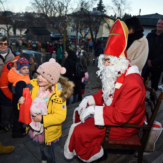 Ein Mann als Weihnachtsmann verkleidet sitzt auf einem Stuhl, während ein junges Mädchen in einer gelben Jacke einen rosa Plüschtier hält.