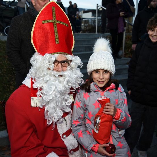 Ein Mann, der als St. Nikolaus verkleidet ist, mit weißem Bart und rotem Hut, steht neben einem jungen Mädchen, das eine weiße Mütze trägt und einen Apfel hält. Hinter ihnen sind weitere Menschen zu sehen, darunter ein lächelnder Junge.