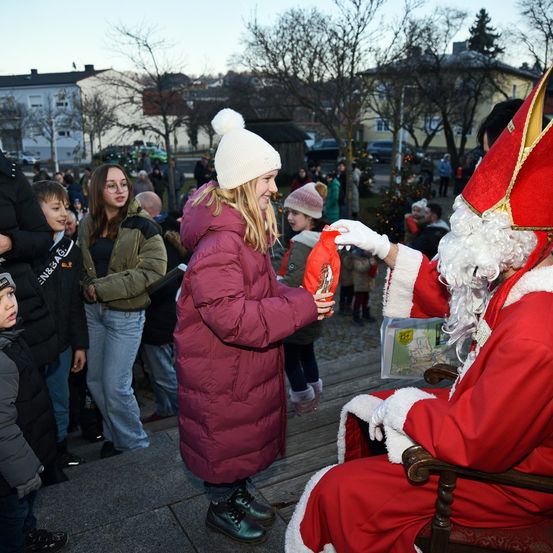 Eine Menschenmenge versammelt sich um den auf einem Stuhl sitzenden Weihnachtsmann, der einem Mädchen ein Geschenk überreicht. Dahinter stehen und gehen Menschen auf der Straße, während Bäume und Gebäude im Hintergrund stehen.