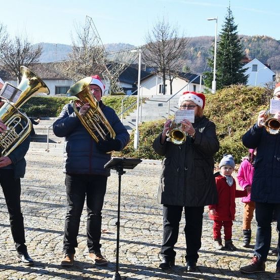 Eine Brassband in Weihnachtsmützen spielt im Freien, begleitet von Kindern und Erwachsenen, die zuschauen.
