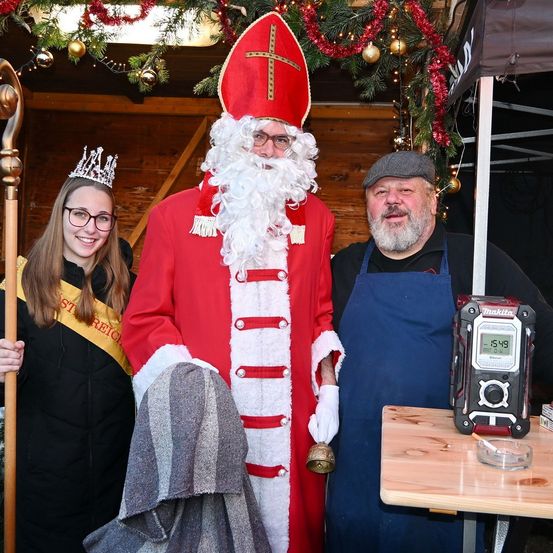 Eine Frau mit Krone und ein Mann in Weihnachtsmannkostüm stehen neben einem Mann in blauer Schürze. Dahinter steht ein Tisch mit Radio und Zigarette. Dahinter steht ein Zelt.