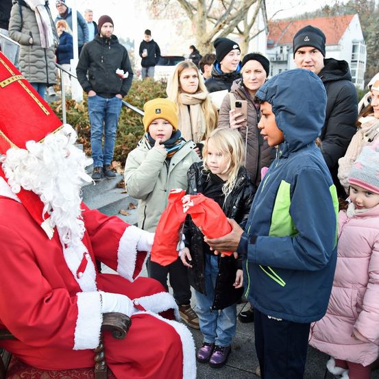 Der Weihnachtsmann in rotem Gewand sitzt auf einem Stuhl und gibt einem jungen Jungen einen roten Beutel, umgeben von anderen Kindern und Erwachsenen. Einige schauen auf ihre Handys.
