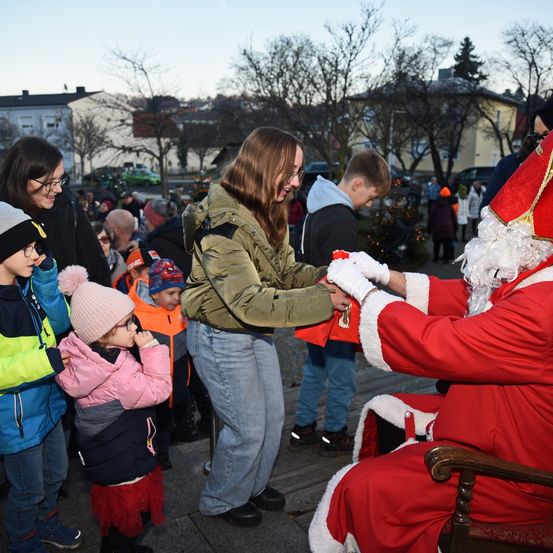 Ein in Rot gekleideter Weihnachtsmann interagiert mit Kindern und verteilt Geschenke. Eine Frau in einer grünen Jacke und Jeans steht neben ihm und lächelt. Die Kinder tragen Winterkleidung und Mützen, und einige halten Geschenke. Im Hintergrund befinden sich Gebäude und Bäume.