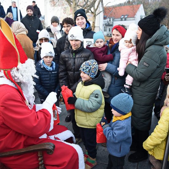 Der Weihnachtsmann in einem roten Kostüm begrüßt Kinder und deren Eltern im öffentlichen Raum. Viele halten rote Taschen.
