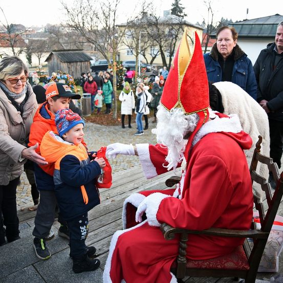 Eine Santa-Claus-Figur, gekleidet in Rot und Weiß, sitzt auf einem Stuhl und überreicht einem kleinen Jungen ein Geschenk vor einer Menschenmenge, darunter eine Frau mit Brille.