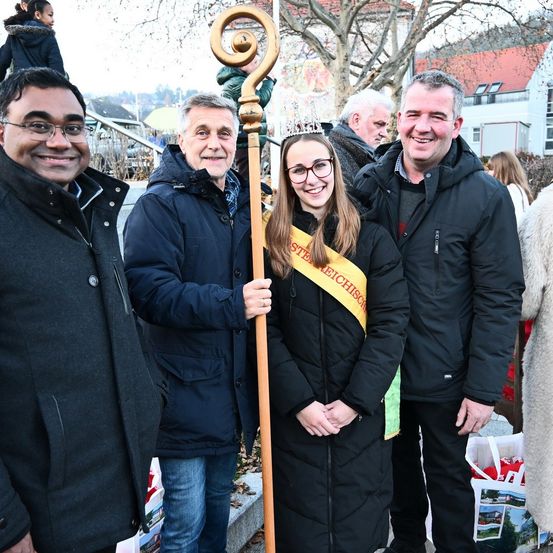 Eine Gruppe von Menschen steht lächelnd zusammen draußen. Eine Frau in der Mitte trägt eine Krone und hält einen Stab. Der Mann links trägt eine Brille. Der Mann rechts hält eine Tasche.