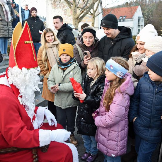 Der Weihnachtsmann spricht mit einer Gruppe von Kindern. Er ist in einem traditionellen roten und weißen Weihnachtsmann-Kostüm gekleidet. Die Kinder halten rote Taschen und lauschen aufmerksam.