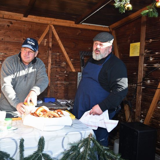 Zwei Männer stehen in einem Holzraum. Einer hält einen Krug und der andere eine Serviette. Sie stehen vor einem Tisch mit Brezeln.