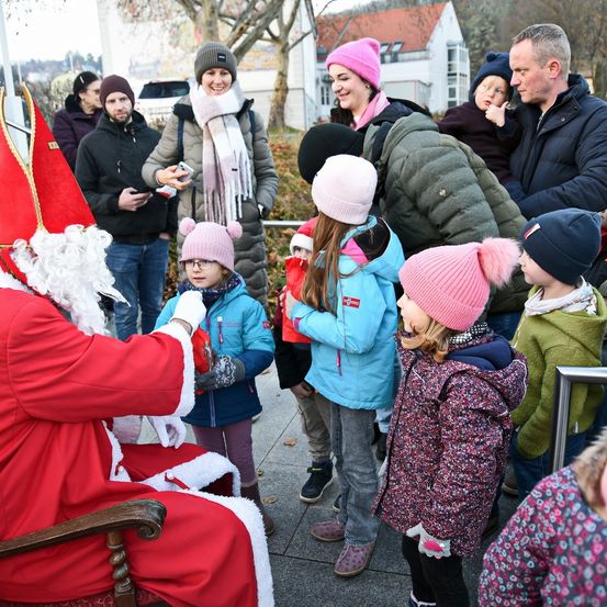 Der Weihnachtsmann begrüßt eine Gruppe von Kindern, umgeben von Erwachsenen, einschließlich Eltern und anderen Familien, in einer festlichen Außenumgebung.