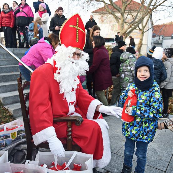 Ein Mann, der als Weihnachtsmann verkleidet ist, sitzt auf einem Stuhl mit einem Kind vor ihm. Sie sind von mehreren Menschen umgeben, die auf Treppen und am Boden stehen.