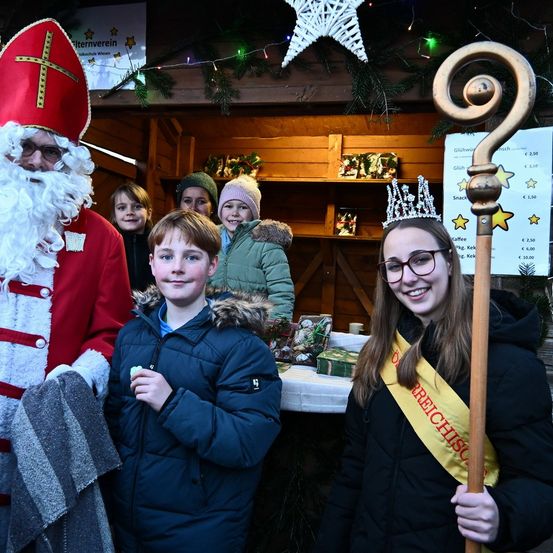 Eine Gruppe von Kindern, zwei Jungen und ein Mädchen, gekleidet in Winterkleidung, steht neben dem Weihnachtsmann in einem Weihnachtsmarktstand.