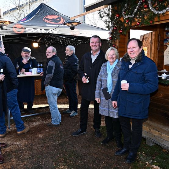 Eine Gruppe von Menschen steht vor einem Zelt mit Weihnachtsdekoration. Ein Mann hält ein Bier und lächelt, während andere plaudern und Tassen halten.