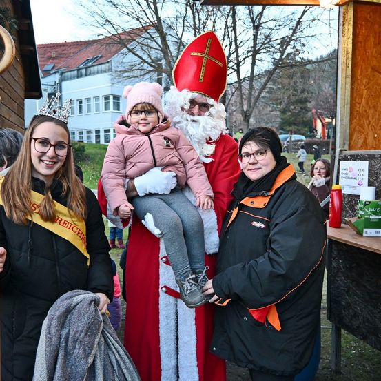 Eine lächelnde Frau mit einer Schärpe, ein Kind mit Brille und ein Mann, der als Weihnachtsmann gekleidet ist und ein Kind hält, posieren für ein Foto draußen.
