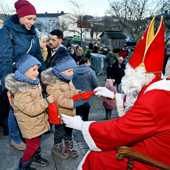 Zwei Kinder erhalten Geschenke von Santa Claus, begleitet von ihren Eltern und anderen Menschen, in einer öffentlichen Umgebung.