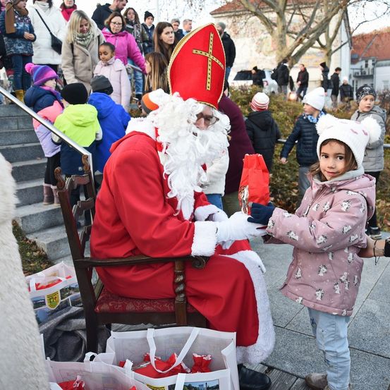 Ein Mann in Santa-Claus-Kleidung, der sitzt, übergibt einem jungen Mädchen ein Geschenk auf einer Stadtstraße mit Zuschauern im Hintergrund.