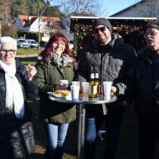 Vier Erwachsene stehen draußen in Winterkleidung. Zwei Männer tragen Mützen und eine Frau trägt eine Brille. Sie lächeln zur Kamera. Ein Tisch mit Essen und Getränken steht zwischen ihnen.