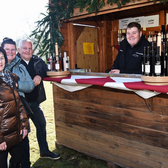 Drei Personen stehen hinter einem Holztresen mit einem Mann dahinter. Flaschen mit Wein und Olivenöl sind auf dem Tresen ausgestellt.
