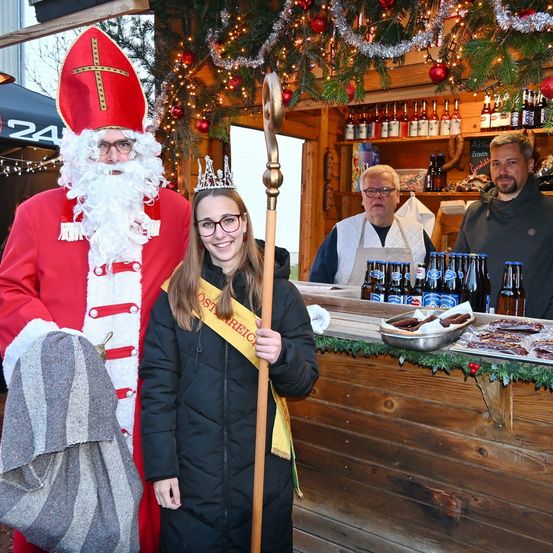 Eine festliche Szene mit einer Frau in einem schwarzen Mantel, die eine gelbe Schärpe und eine Krone trägt, neben einem Mann, der als Weihnachtsmann verkleidet ist. Hinter ihnen stehen zwei Männer an einer Bartheke.