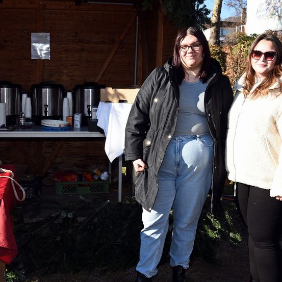 Zwei Frauen stehen vor einem Holzschuppen mit einem Tisch. Die Frau auf der linken Seite trägt eine Brille und eine Jacke. Hinter ihnen befinden sich mehrere Behälter und ein Haufen Kiefernzweige.