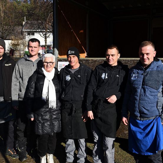 Eine Gruppe von sechs Personen steht zusammen draußen. Sie tragen Winterkleidung. Eine Frau trägt eine Brille und einen Schal. Hinter ihnen steht eine Holzkonstruktion.