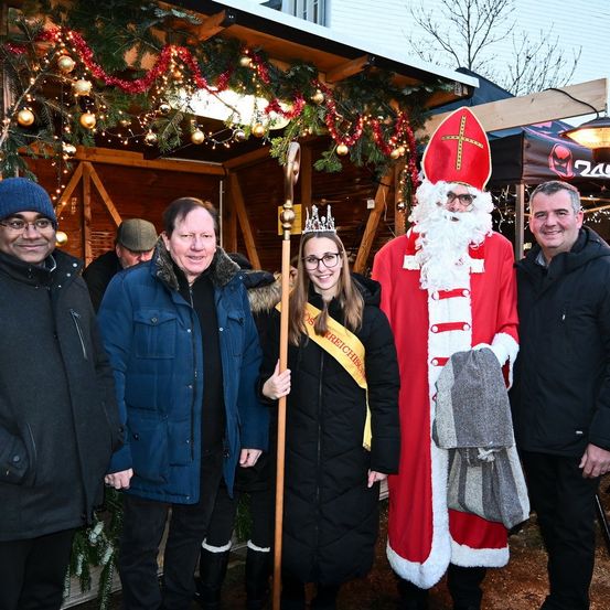 Eine Gruppe von Menschen posiert für ein Foto vor einem festlich geschmückten Stand, wobei eine Frau eine Krone trägt und ein Mann als Weihnachtsmann gekleidet ist. Hinter ihnen befinden sich Weihnachtsdekorationen und ein Zelt.