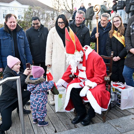 Eine Gruppe von Menschen versammelt sich draußen mit einem als Weihnachtsmann verkleideten Mann, der auf einem Stuhl sitzt und mit einem kleinen Mädchen spricht.
