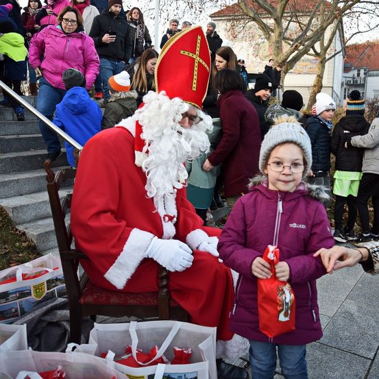 Eine Person, die als Weihnachtsmann verkleidet ist, sitzt auf einem Stuhl, während ein kleines Mädchen in einer lila Jacke neben ihm steht. Hinter ihnen versammeln sich mehrere Personen, einige tragen Mützen und Handschuhe. Taschen und Gegenstände liegen auf dem Boden in der Nähe.