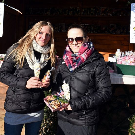 Zwei Frauen stehen in einer gemütlichen Umgebung im Innenbereich, beide in Winterkleidung. Eine Frau hält eine Box mit Snacks und die andere eine Tüte. Hinter ihnen steht ein Tisch mit einer weißen Tischdecke.