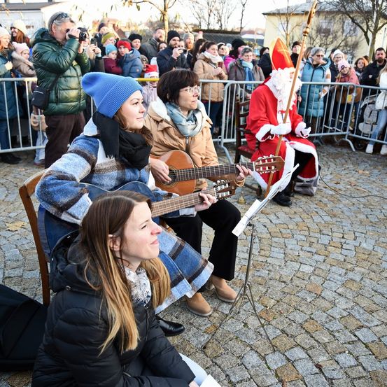 Zwei Frauen spielen Gitarren, während sie auf einem Kopfsteinpflasterplatz sitzen. Eine Menschenmenge beobachtet, darunter ein Mann in Santa-Claus-Kleidung.