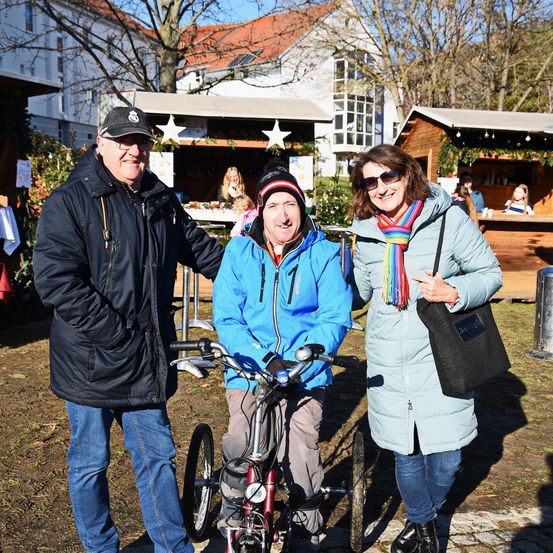 Drei Personen stehen draußen in der Nähe eines Holzhäuschens. Der Mann links trägt eine Mütze und eine Brille, und der Mann in der Mitte trägt eine Mütze. Die Frau rechts lächelt und trägt eine Sonnenbrille.