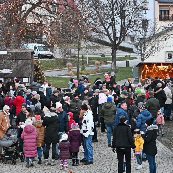 Eine große Menschenmenge versammelt sich im Freien in der Nähe eines Weihnachtsbaums, viele in Winterkleidung. Der Ort scheint ein öffentlicher Platz zu sein, mit Gebäuden und geparkten Fahrzeugen im Hintergrund. Einige Personen halten Kinder, und die Atmosphäre ist festlich.