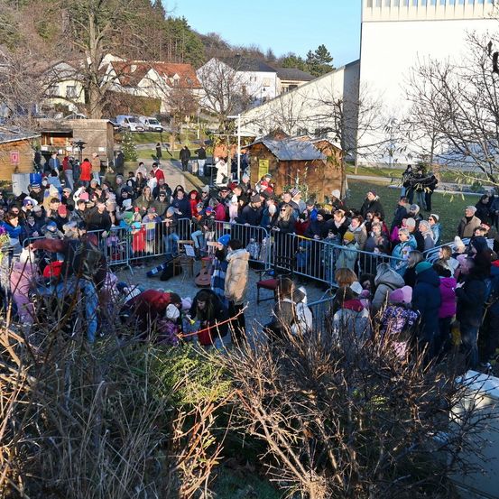 Eine große Menschenmenge, in warme Kleidung gehüllt, versammelt sich im Freien. Sie stehen hinter Metallbarrieren, möglicherweise für eine festliche Veranstaltung. Mehrere kleine Hütten sind verstreut, und im Hintergrund befinden sich Gebäude und Bäume.