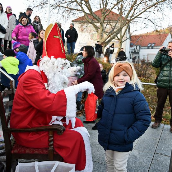 Ein Mann, der als Weihnachtsmann verkleidet ist, sitzt draußen auf einem Stuhl, hält einen roten Beutel und interagiert mit einem jungen Mädchen in einer blauen Jacke. Dahinter stehen mehrere Personen, und eine macht ein Foto. Gebäude sind im Hintergrund.