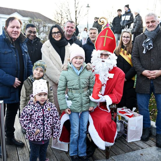 Eine Gruppe von Menschen steht auf einer Holzterrasse. In der Mitte ist eine Person als Weihnachtsmann verkleidet. Einige Leute lächeln und halten Geschenke. Dahinter scheint eine Menschenmenge zuzusehen.