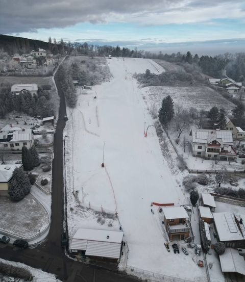 Eine Luftaufnahme einer verschneiten Skipiste mit einer Straße und mehreren Häusern. Das Gebiet ist von Bäumen umgeben, und in der Nähe sind schneebedeckte Autos geparkt.