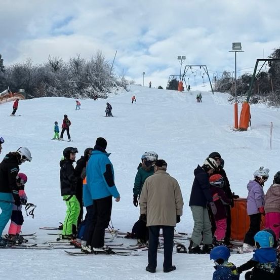 Eine Gruppe von Skifahrern und Snowboardern versammelt sich auf einer verschneiten Piste. Einige tragen Helme, andere halten Skistöcke. Einige Personen fahren die Piste hinunter. Straßenlaternen und orangefarbene Barrieren sind sichtbar.