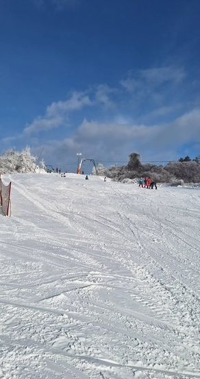 Eine Ski-Piste mit Skifahrern und Snowboardern. Bäume und eine Skilift sind in der Ferne unter teilweise bewölktem Himmel zu sehen.