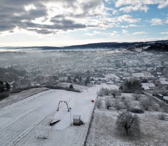 Luftaufnahme einer verschneiten Landschaft mit einer Stadt im Hintergrund, Bergen und einer Skipiste.