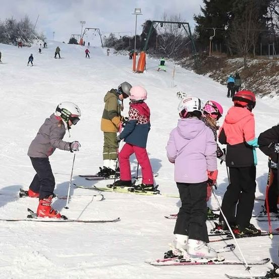 Eine Gruppe von Kindern und Erwachsenen skifahren auf einem verschneiten Hang, tragen Winterkleidung und Helme. Bäume und eine Skilift sind im Hintergrund.
