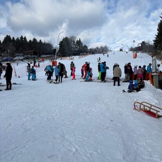 Eine Gruppe von Menschen auf einem verschneiten Hang, einige sind beim Skifahren, andere stehen. Bäume und ein bewölkter Himmel im Hintergrund.