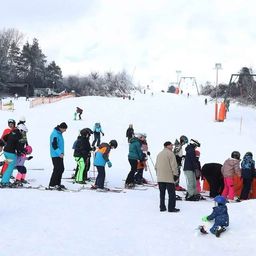 Eine Gruppe von Menschen, einschließlich Kindern, Ski auf einem verschneiten Berg. Einige stehen, andere sitzen. Bäume und eine Skilift im Hintergrund.